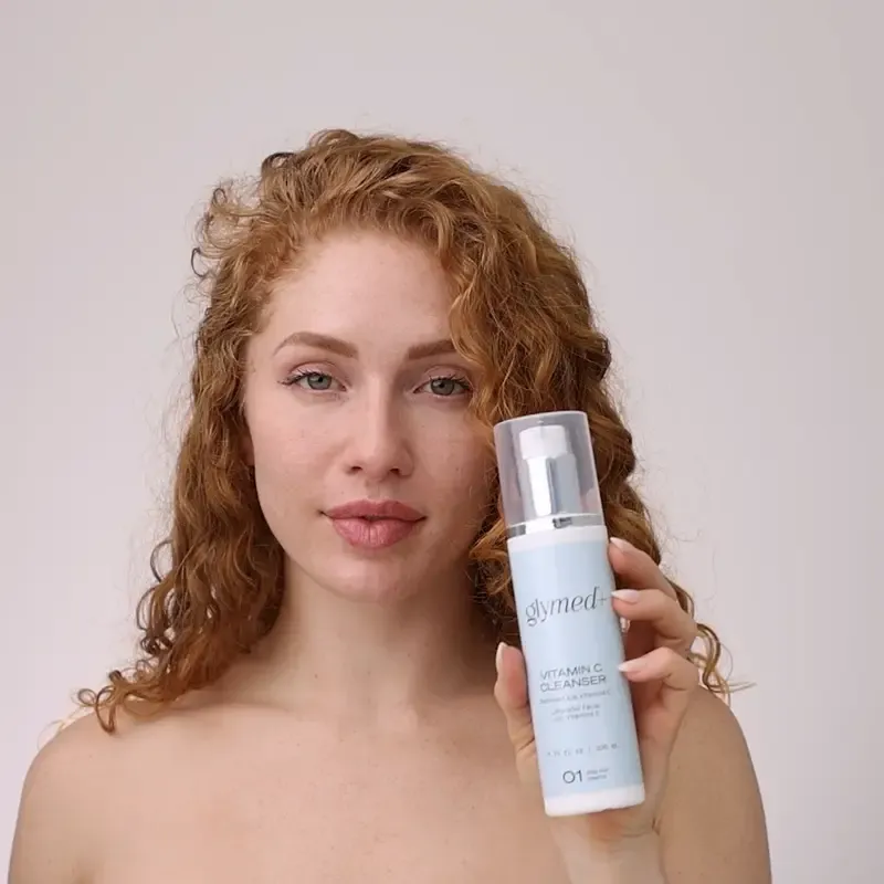 Redhead woman with curly hair holding a white and blue skincare product bottle.